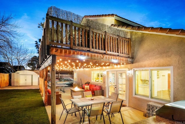 back house at dusk with a balcony, a patio area, a lawn, and a storage unit