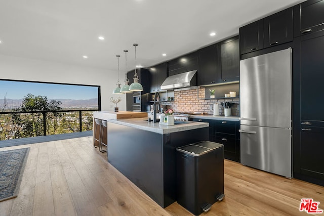 kitchen with an island with sink, light wood-type flooring, wall chimney range hood, pendant lighting, and stainless steel refrigerator