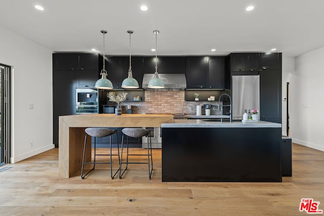 kitchen featuring pendant lighting, appliances with stainless steel finishes, exhaust hood, light hardwood / wood-style floors, and a center island with sink