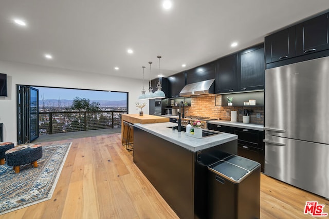 kitchen with wall chimney range hood, pendant lighting, light wood-type flooring, a kitchen island with sink, and stainless steel fridge