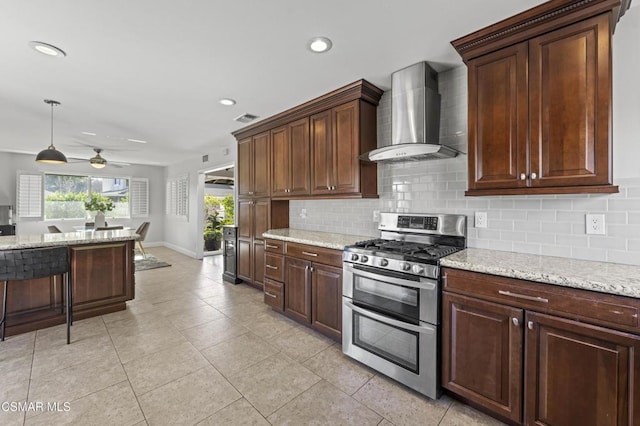 kitchen featuring light stone countertops, wall chimney exhaust hood, range with two ovens, hanging light fixtures, and ceiling fan