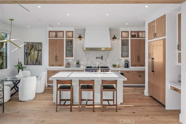 kitchen featuring light wood-type flooring, custom exhaust hood, backsplash, and a kitchen island with sink