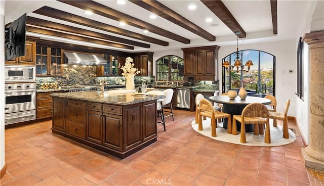 kitchen featuring appliances with stainless steel finishes, a center island with sink, tasteful backsplash, and wall chimney range hood