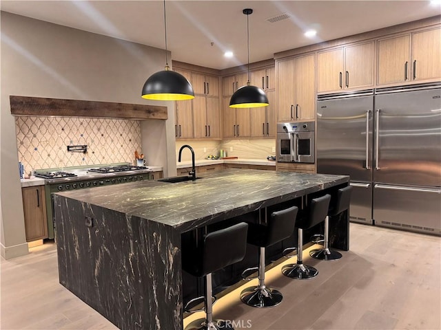 kitchen with stainless steel appliances, a sink, hanging light fixtures, dark stone counters, and a center island with sink