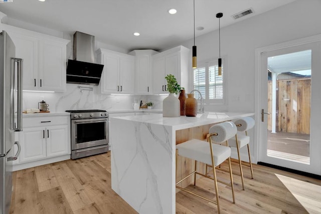 kitchen featuring pendant lighting, white cabinetry, light stone counters, stainless steel appliances, and wall chimney range hood