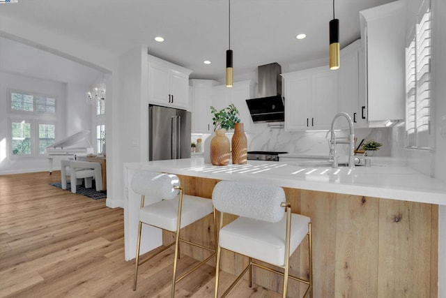 kitchen with wall chimney range hood, high end fridge, hanging light fixtures, white cabinetry, and a kitchen breakfast bar