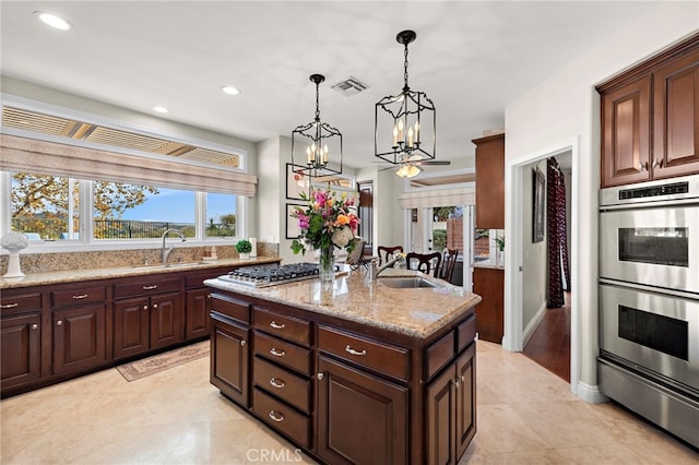 kitchen with pendant lighting, appliances with stainless steel finishes, sink, light stone counters, and a center island with sink