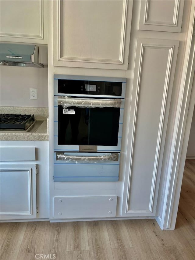 kitchen featuring black gas cooktop, stainless steel oven, wall chimney exhaust hood, light stone countertops, and light hardwood / wood-style flooring