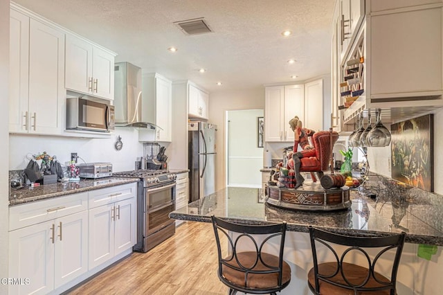 kitchen with wall chimney range hood, appliances with stainless steel finishes, a breakfast bar area, and dark stone countertops