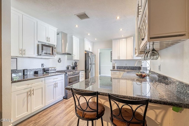 kitchen featuring white cabinets, wall chimney exhaust hood, a breakfast bar, dark stone countertops, and stainless steel appliances