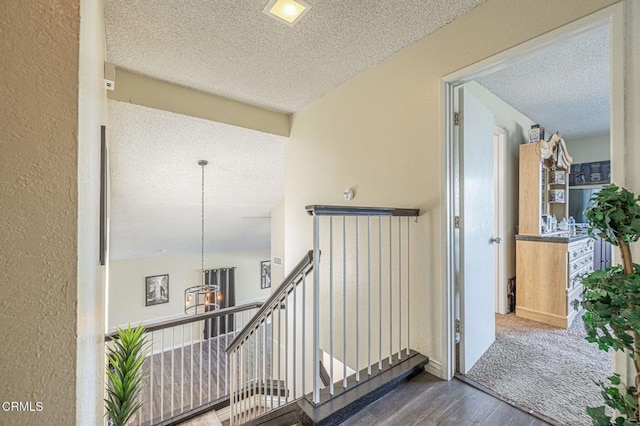 hallway with a textured ceiling, a textured wall, dark wood-style flooring, and an upstairs landing