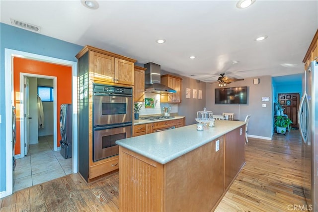 kitchen featuring visible vents, light wood-style flooring, a kitchen island, appliances with stainless steel finishes, and wall chimney range hood