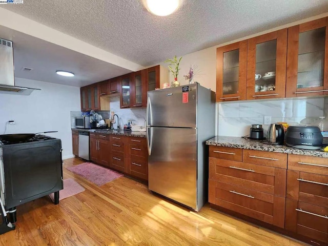 kitchen with appliances with stainless steel finishes, tasteful backsplash, stone countertops, wall chimney exhaust hood, and light wood-type flooring