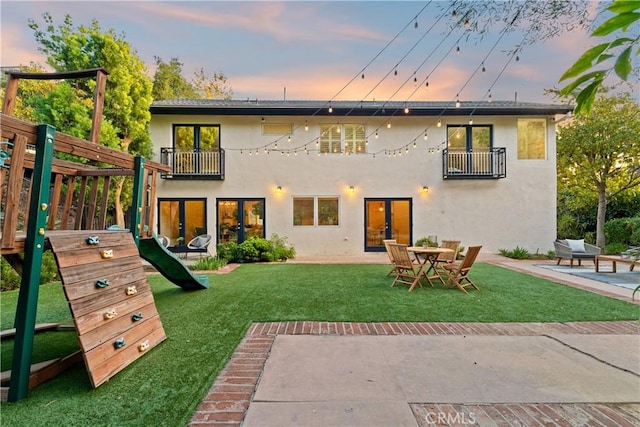 back house at dusk featuring a lawn, a patio area, a playground, french doors, and a balcony