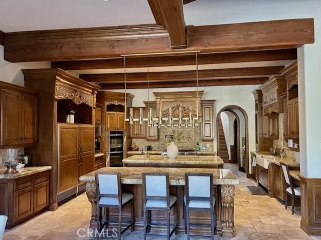 kitchen featuring decorative light fixtures, beam ceiling, a breakfast bar area, a center island with sink, and double oven