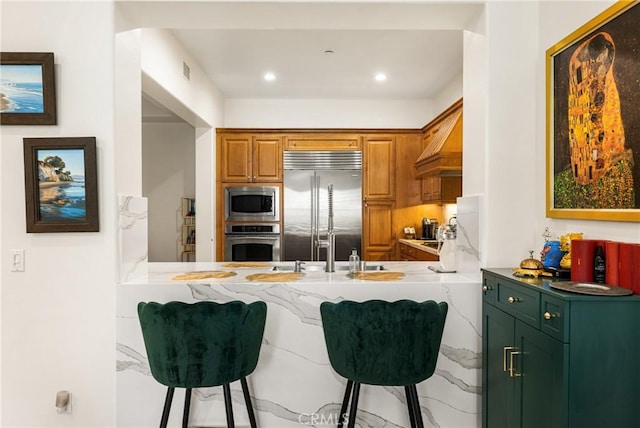 kitchen featuring premium range hood, a kitchen bar, kitchen peninsula, and built in appliances