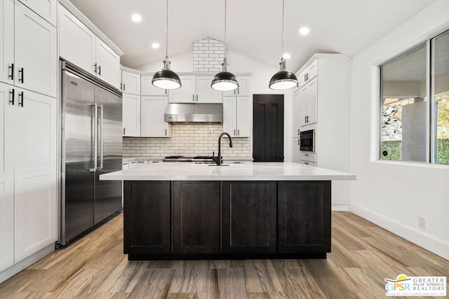 kitchen with pendant lighting, a kitchen island with sink, vaulted ceiling, built in fridge, and light wood-type flooring