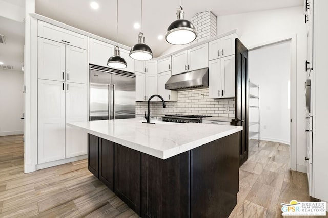 kitchen with pendant lighting, built in refrigerator, light stone counters, an island with sink, and white cabinets