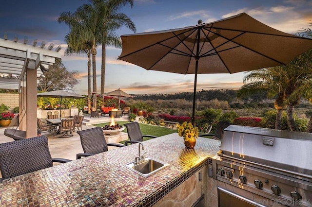 patio terrace at dusk featuring a fire pit, an outdoor wet bar, exterior kitchen, grilling area, and a pergola