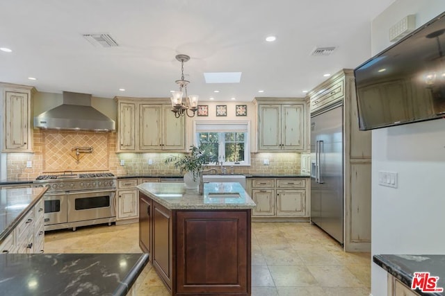 kitchen featuring high quality appliances, pendant lighting, wall chimney range hood, and cream cabinets