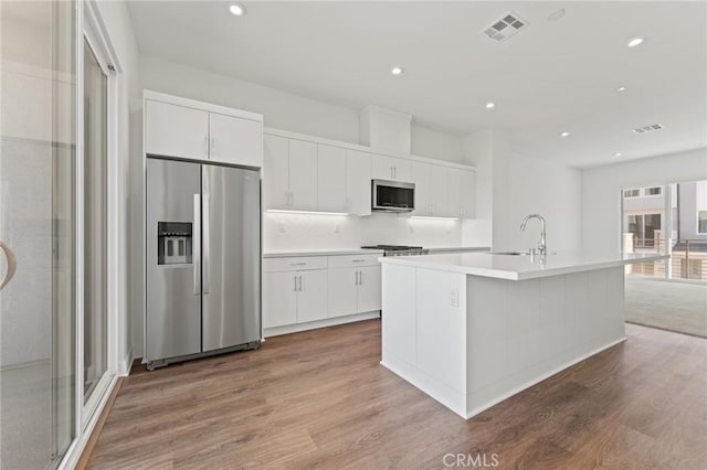 kitchen featuring wood-type flooring, sink, a kitchen island with sink, appliances with stainless steel finishes, and white cabinets