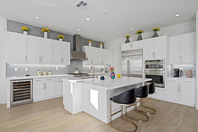 kitchen with white cabinets, beverage cooler, wall chimney exhaust hood, stainless steel appliances, and an island with sink