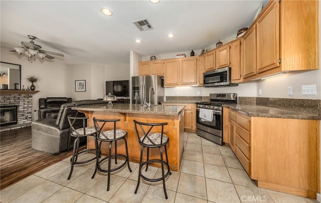 kitchen featuring light tile patterned floors, ceiling fan, stainless steel appliances, a brick fireplace, and a kitchen island with sink