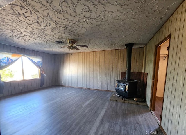 unfurnished living room with wood walls, ceiling fan, a wood stove, dark hardwood / wood-style floors, and a textured ceiling