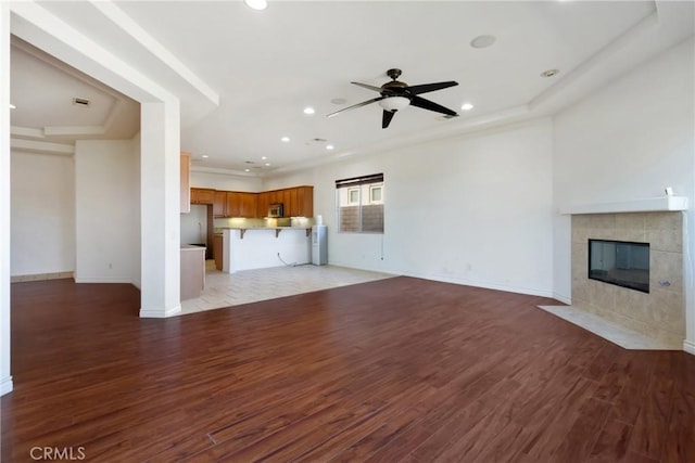 unfurnished living room with ceiling fan, a tiled fireplace, a raised ceiling, and light hardwood / wood-style floors