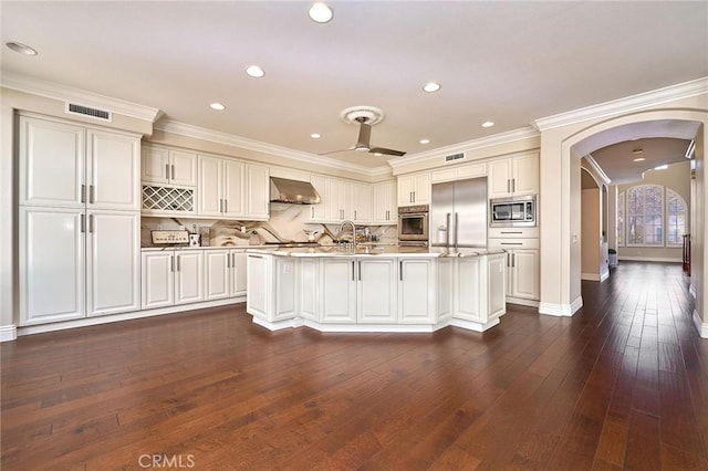 kitchen featuring decorative backsplash, wall chimney exhaust hood, built in appliances, and an island with sink