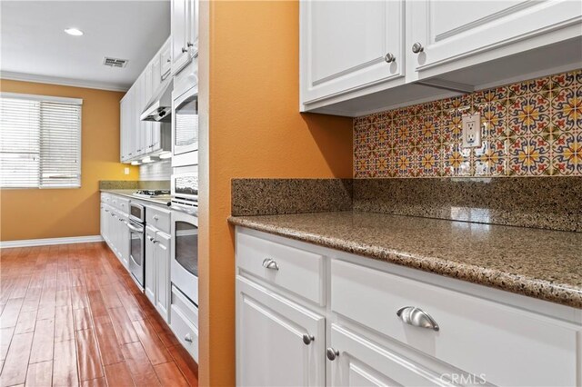 kitchen with stone countertops, wood-type flooring, white cabinets, backsplash, and stainless steel appliances