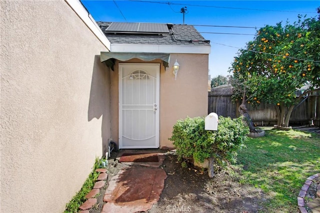 property entrance featuring a shingled roof, roof mounted solar panels, fence, and stucco siding