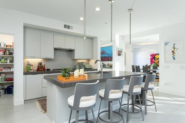 kitchen with light tile patterned floors, an island with sink, gray cabinetry, decorative backsplash, and sink