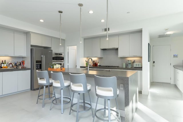 kitchen with a center island with sink, pendant lighting, decorative backsplash, gray cabinetry, and stainless steel appliances