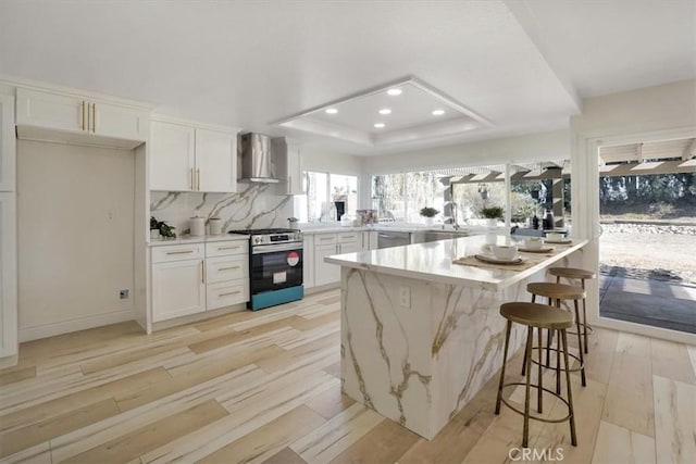 kitchen with wall chimney range hood, a raised ceiling, appliances with stainless steel finishes, a breakfast bar area, and white cabinets