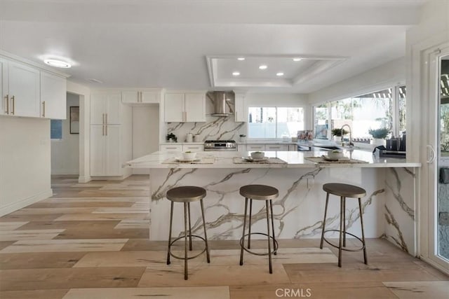 kitchen with white cabinetry, a kitchen bar, a tray ceiling, stove, and wall chimney exhaust hood