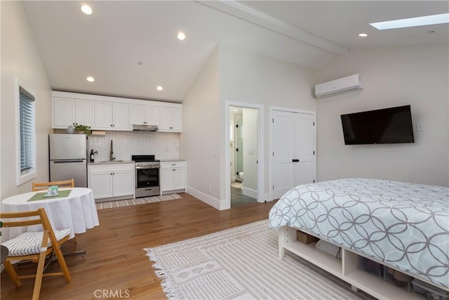 bedroom featuring connected bathroom, lofted ceiling with skylight, a wall mounted AC, stainless steel refrigerator, and light hardwood / wood-style floors