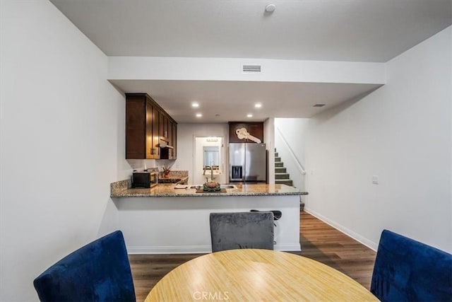 kitchen featuring dark wood-type flooring, stone countertops, stainless steel fridge with ice dispenser, kitchen peninsula, and dark brown cabinets