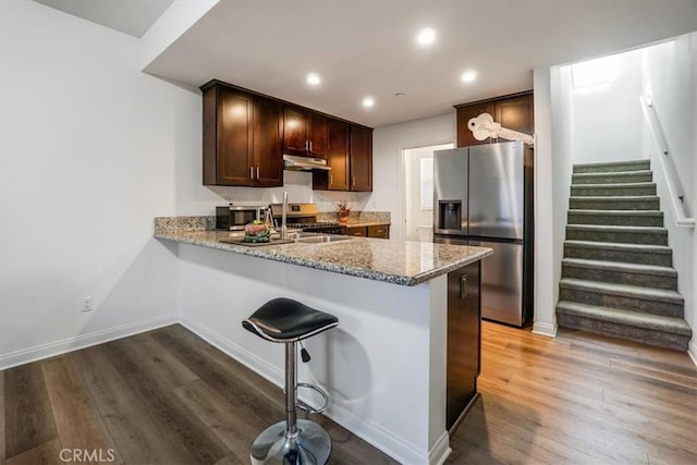 kitchen featuring kitchen peninsula, dark brown cabinets, appliances with stainless steel finishes, dark hardwood / wood-style flooring, and light stone counters