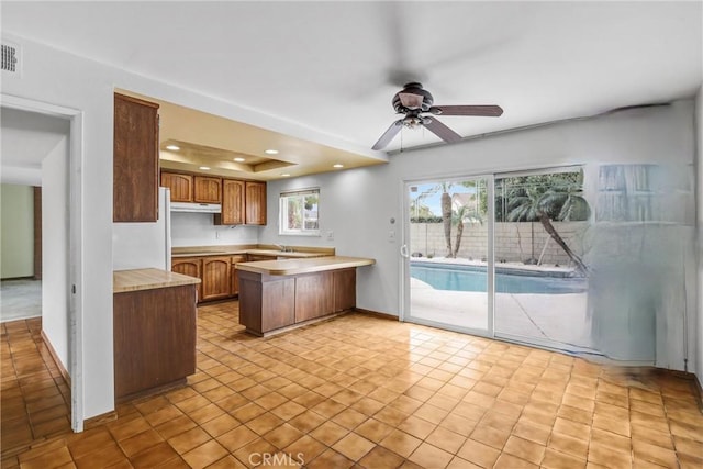 kitchen featuring brown cabinets, a raised ceiling, light countertops, under cabinet range hood, and a peninsula