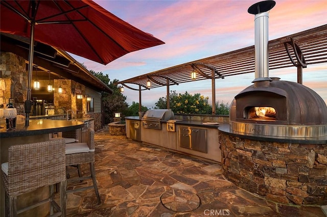 patio terrace at dusk with exterior kitchen, a pergola, a grill, and exterior fireplace