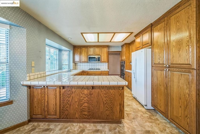 kitchen with tile countertops, kitchen peninsula, gas cooktop, backsplash, and white fridge with ice dispenser