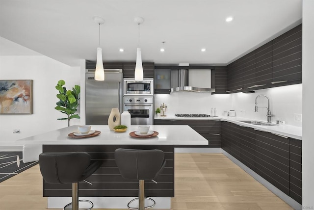 kitchen featuring pendant lighting, wall chimney range hood, built in appliances, sink, and a kitchen breakfast bar