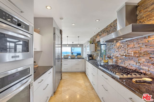 kitchen featuring white cabinetry, stainless steel appliances, dark stone counters, wall chimney range hood, and sink