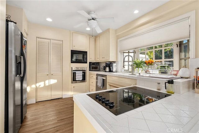 kitchen featuring ceiling fan, black appliances, light wood-type flooring, tile counters, and cream cabinets