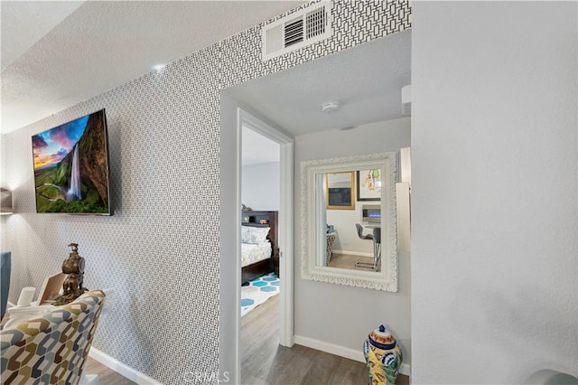 hall featuring visible vents, a textured ceiling, wood finished floors, baseboards, and wallpapered walls