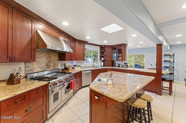 kitchen with a center island with sink, stainless steel appliances, wall chimney range hood, light stone counters, and sink