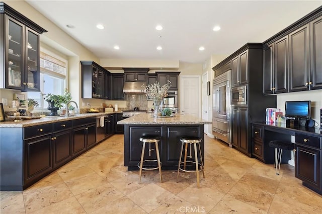 kitchen with built in study area, a kitchen island, light stone counters, built in appliances, and under cabinet range hood