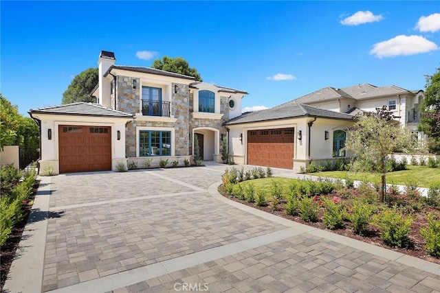 view of front of home with decorative driveway, a chimney, a balcony, a garage, and stone siding