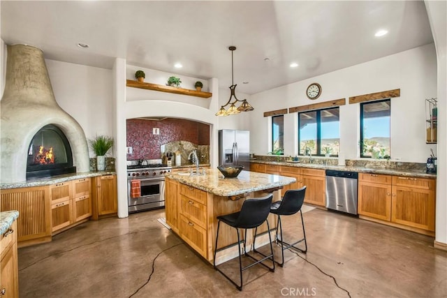 kitchen featuring stainless steel appliances, backsplash, hanging light fixtures, a kitchen island with sink, and light stone counters